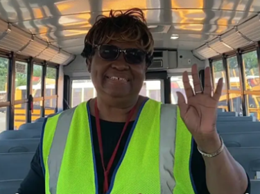  person in a yellow vest standing in a school bus, smiling & waving at the camera